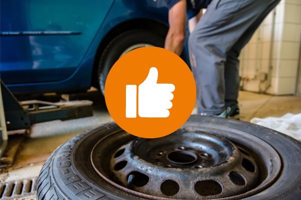 tyre lying on floor of garage with mechanic working in background; a thumbs up graphic in an orange circle is in the centre of the photo - Nut Jobb multipurpose spray